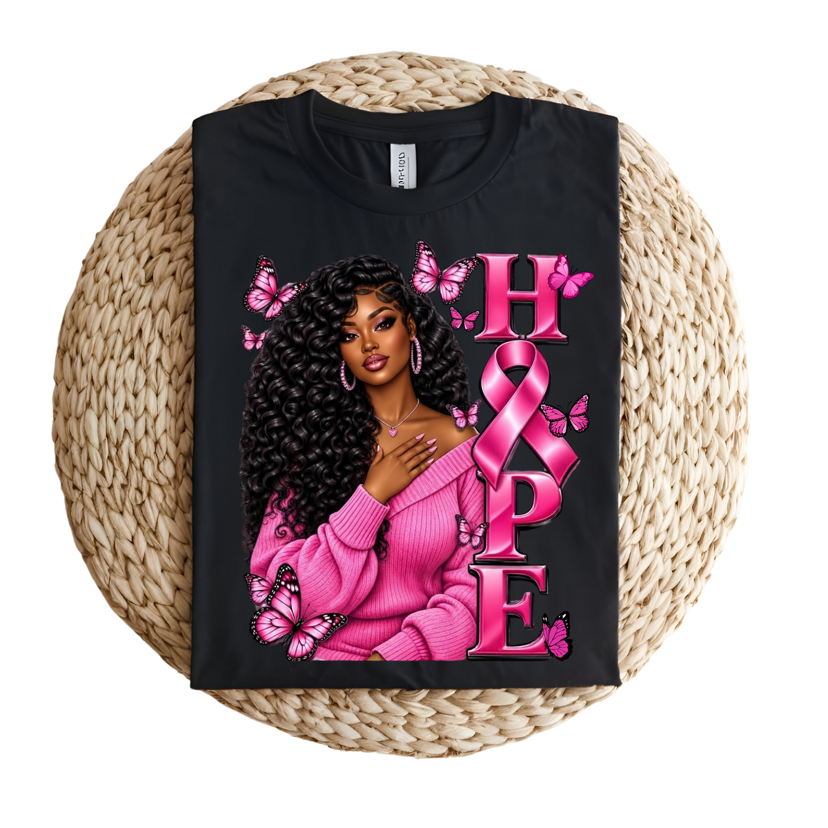 A black short-sleeve T-shirt laid flat on a round woven straw mat, featuring a pink graphic with a ribbon, boxing gloves, and the words 'Breast Cancer WARRIOR' in pink lettering.