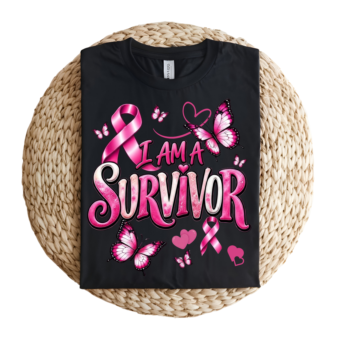 A black short-sleeve T-shirt laid flat on a round woven straw mat, featuring a large pink and white graphic that reads “I AM A SURVIVOR” with pink awareness ribbons, butterflies, and hearts.