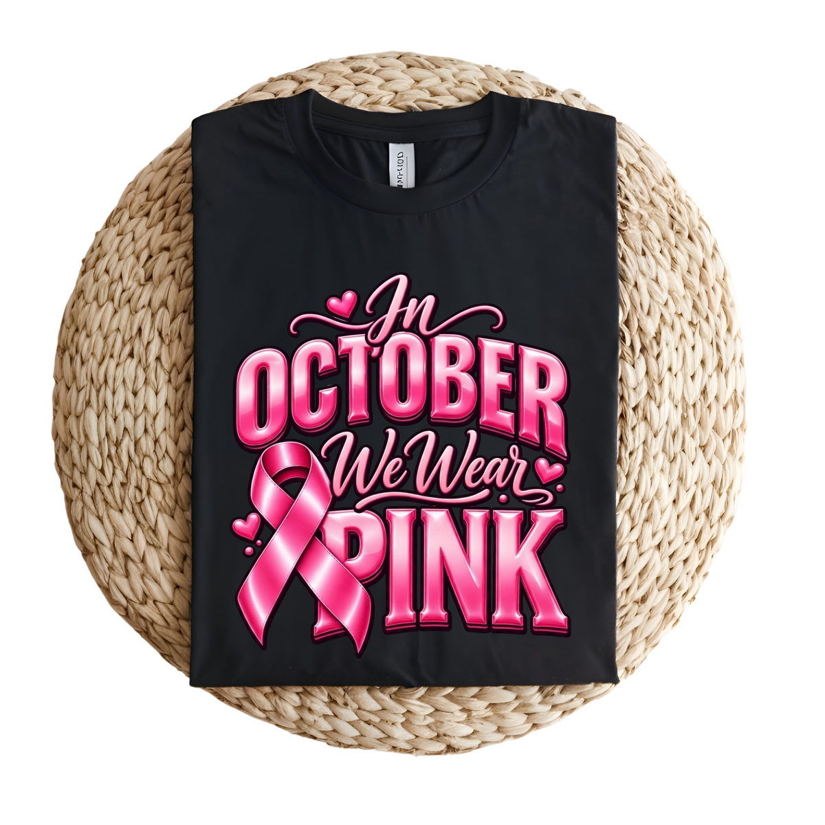 A black short-sleeve T-shirt laid flat on a round woven straw mat, featuring a pink graphic with a ribbon, boxing gloves, and the words 'Breast Cancer WARRIOR' in pink lettering.