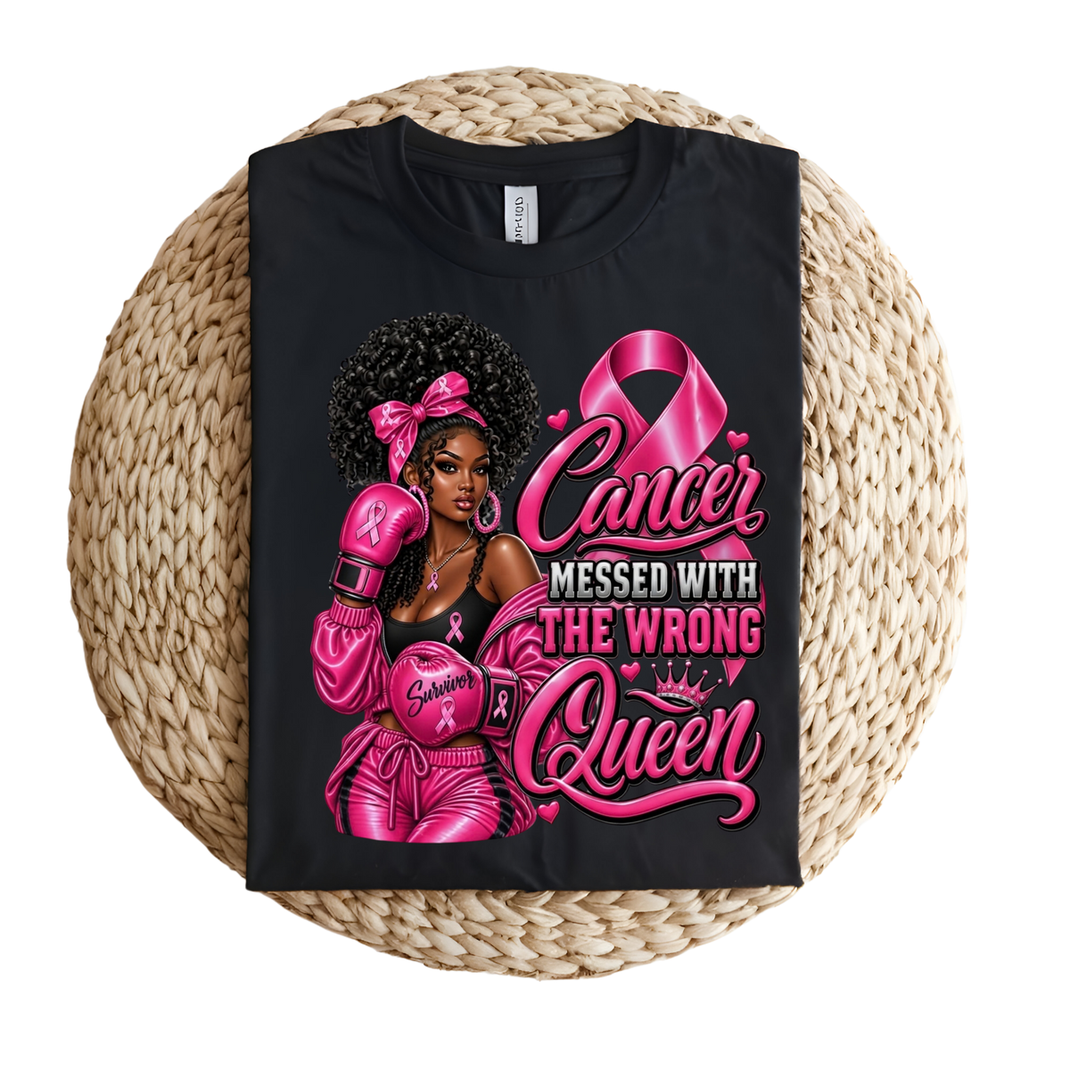 A black short-sleeve graphic T-shirt laid flat on a round woven straw mat. The shirt features a large pink and white graphic of a Black woman with an afro, pink boxing gloves, and the text 'Cancer Messed With The Wrong Queen' alongside a pink ribbon and crown.