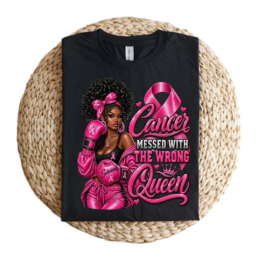 A black short-sleeve graphic T-shirt laid flat on a round woven straw mat. The shirt features a large pink and white graphic of a Black woman with an afro, pink boxing gloves, and the text 'Cancer Messed With The Wrong Queen' alongside a pink ribbon and crown.