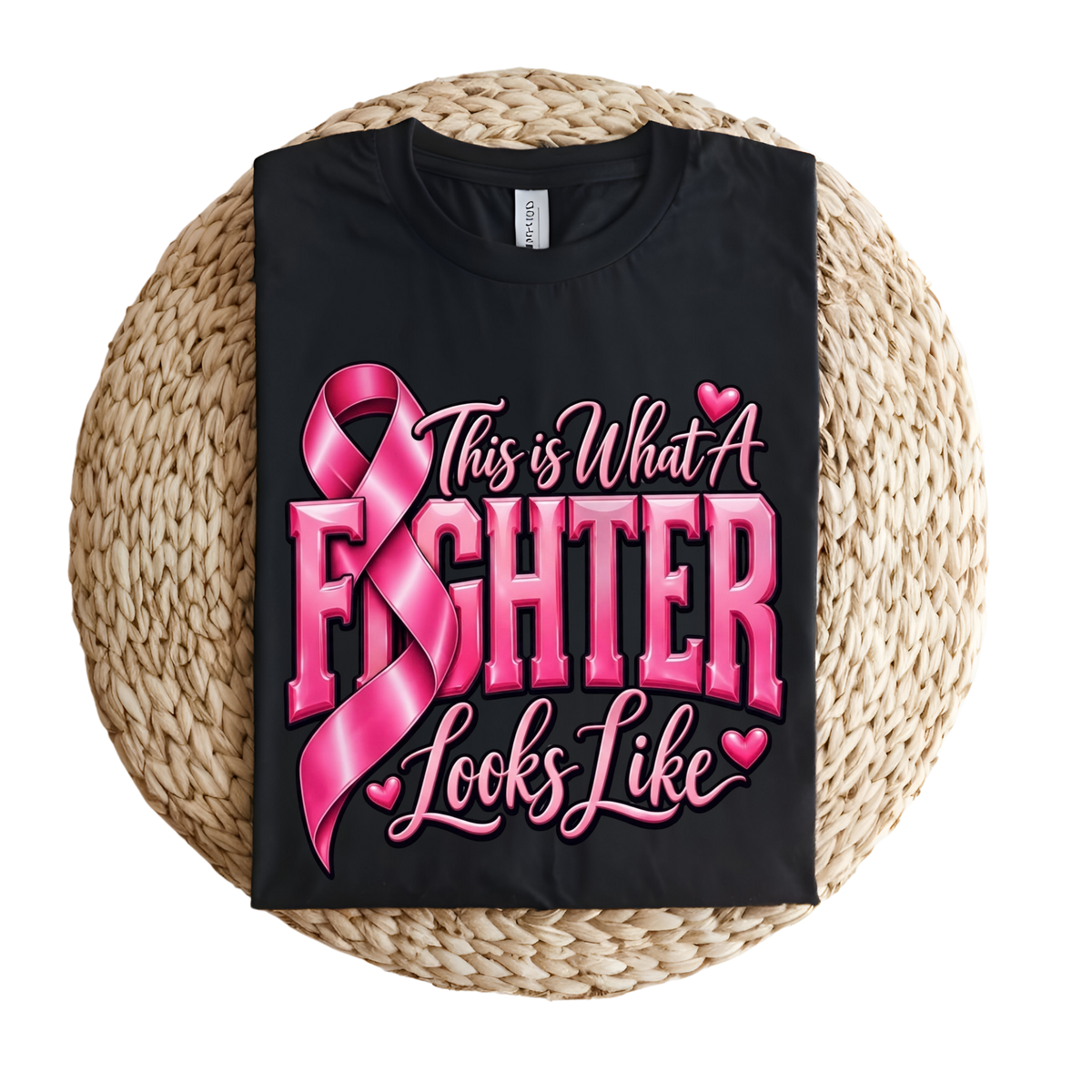 A black short-sleeve T-shirt laid flat on a round woven straw mat, featuring a pink graphic with a ribbon, boxing gloves, and the words 'Breast Cancer WARRIOR' in pink lettering.