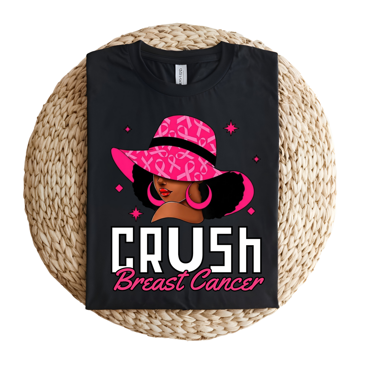 A black short-sleeve T-shirt laid flat on a round woven straw mat, featuring a graphic of a woman in a pink leopard-print hat with the text 'CRUSH Breast Cancer' in white and pink.