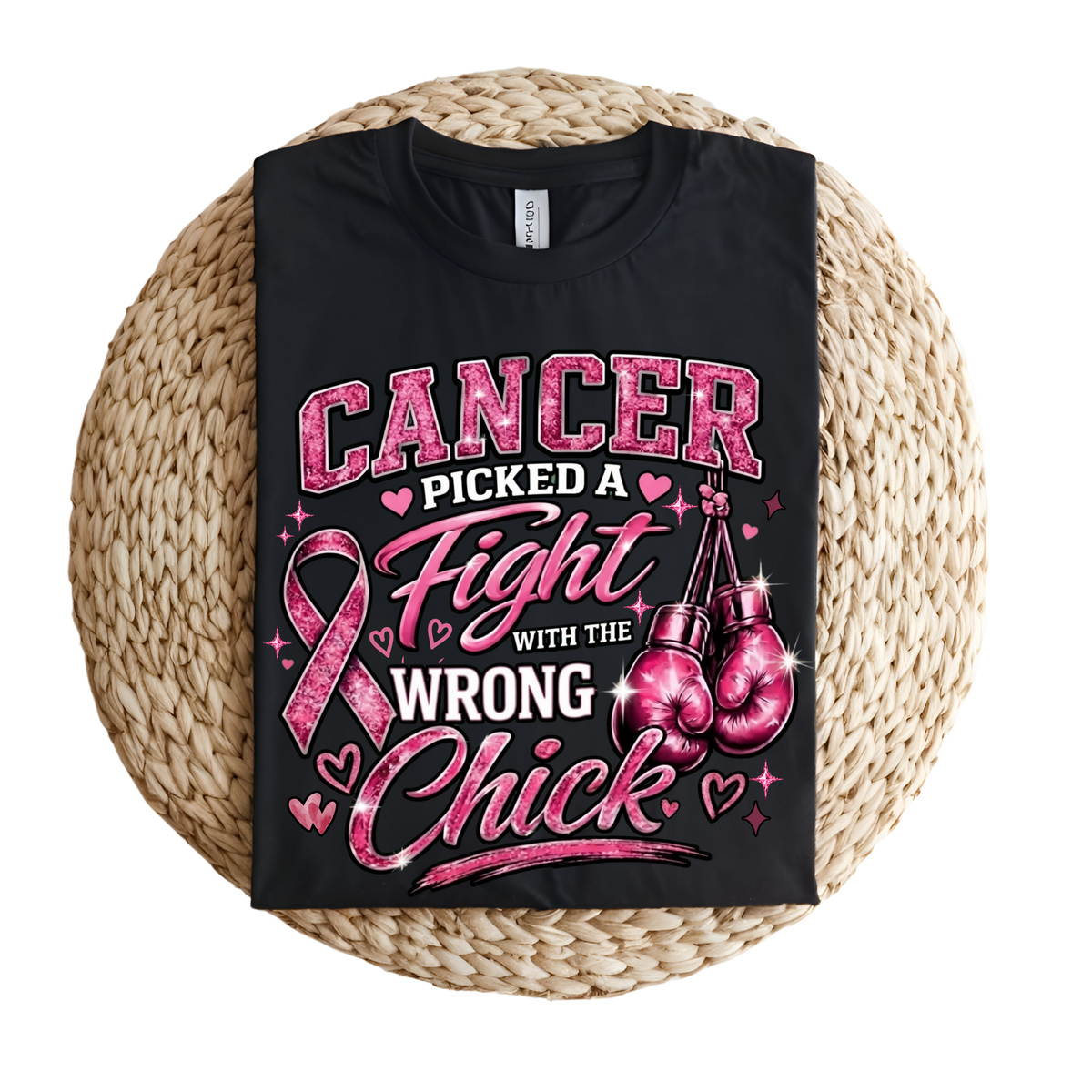 A black short-sleeve T-shirt laid flat on a round woven straw mat, featuring a pink and white graphic that reads “CANCER PICKED A Fight WITH THE WRONG Chick,” with a pink ribbon, hearts, and boxing gloves.
