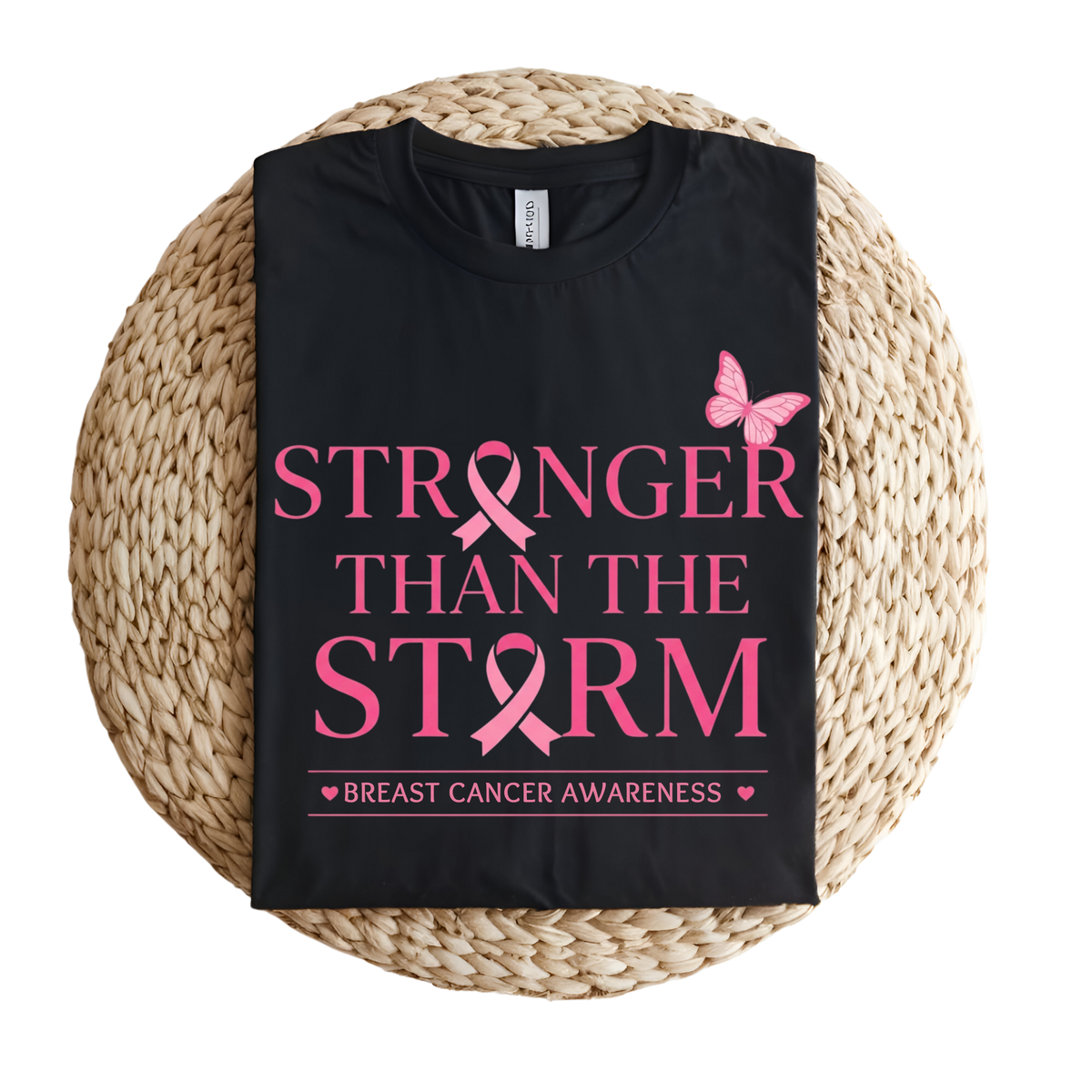 A black short-sleeve T-shirt laid flat on a round woven straw mat, featuring pink text that reads “STRONGER THAN THE STORM” with pink ribbon graphics and a pink butterfly, plus smaller text reading “BREAST CANCER AWARENESS” at the bottom.