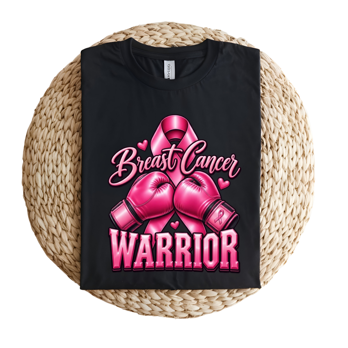 A black short-sleeve T-shirt laid flat on a round woven straw mat, featuring a pink graphic with a ribbon, boxing gloves, and the words 'Breast Cancer WARRIOR' in pink lettering.