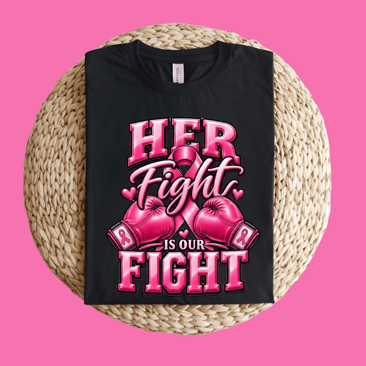 A black short-sleeve T-shirt laid flat on a round woven straw mat against a solid pink background. The shirt features a bold graphic with the words 'HER Fight IS OUR FIGHT' in pink and white, accompanied by pink boxing gloves and a pink ribbon.