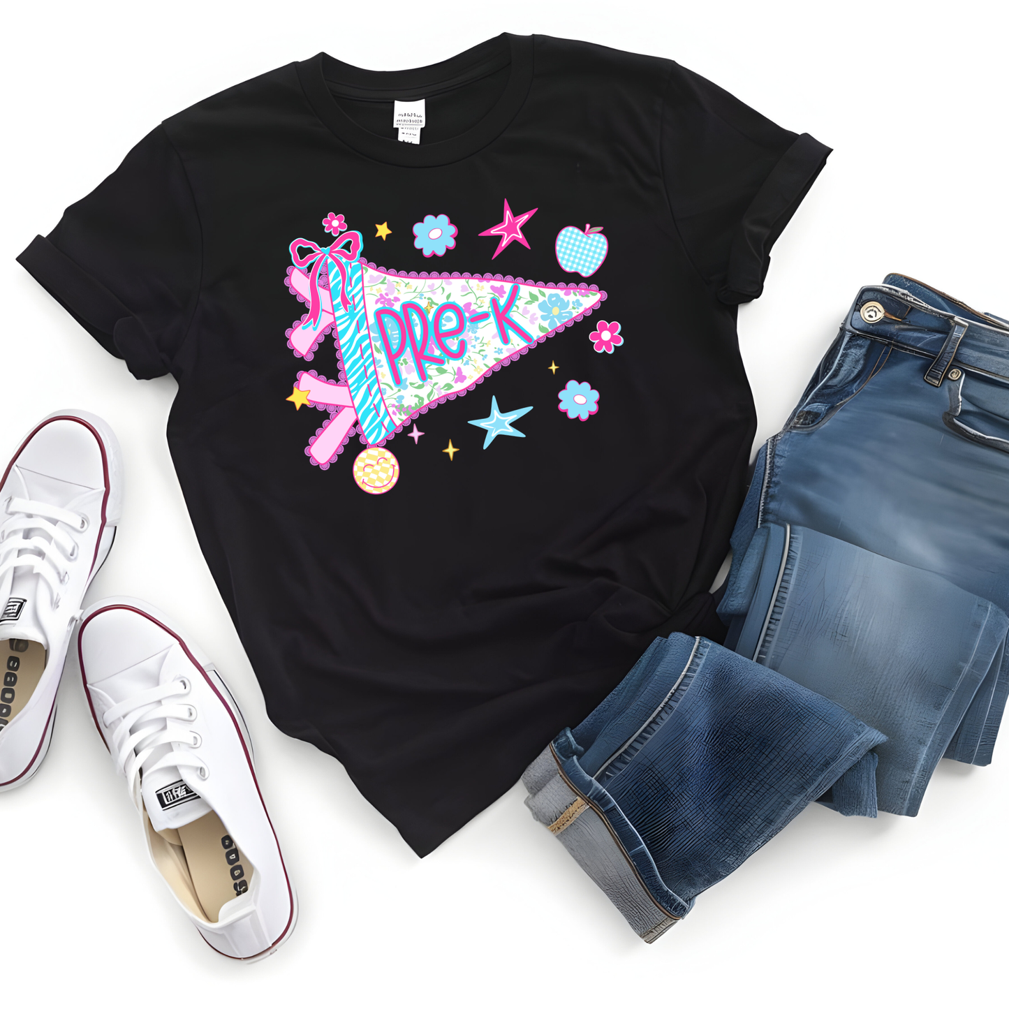 A black short-sleeve graphic T-shirt laid flat on a white background, styled with blue jeans and white sneakers. The shirt features a colorful 'Pre-K' banner design with stars, flowers, and a ribbon motif.