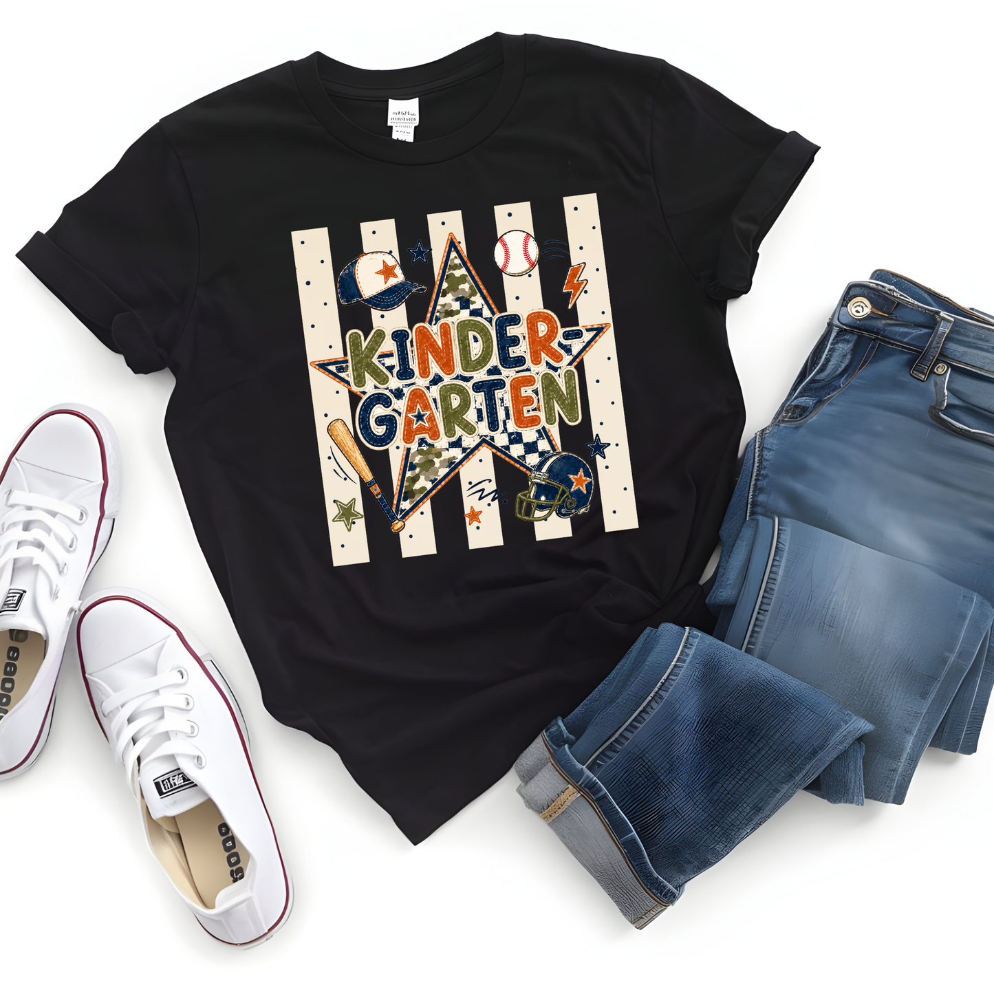 A black short-sleeve graphic T-shirt laid flat with jeans and white sneakers around it on a white background. The shirt has a baseball-themed 'KINDER GARTEN' graphic with stripes, stars, bats, balls, and a helmet.