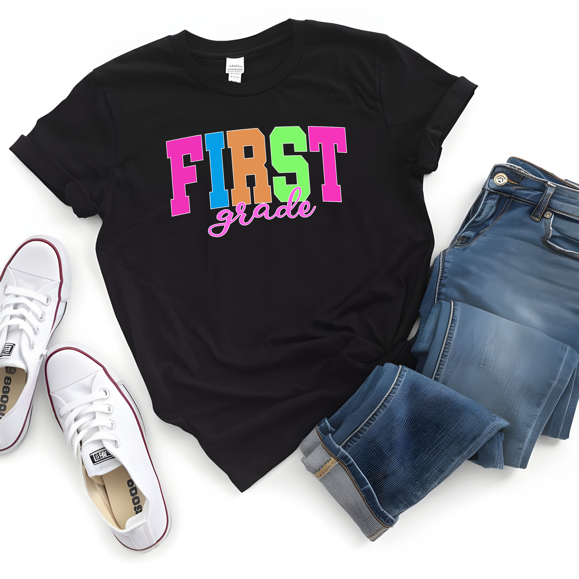 A black graphic t-shirt with 'FIRST grade' printed in colorful letters, styled in a flat lay with blue jeans, white sneakers, and a white background.