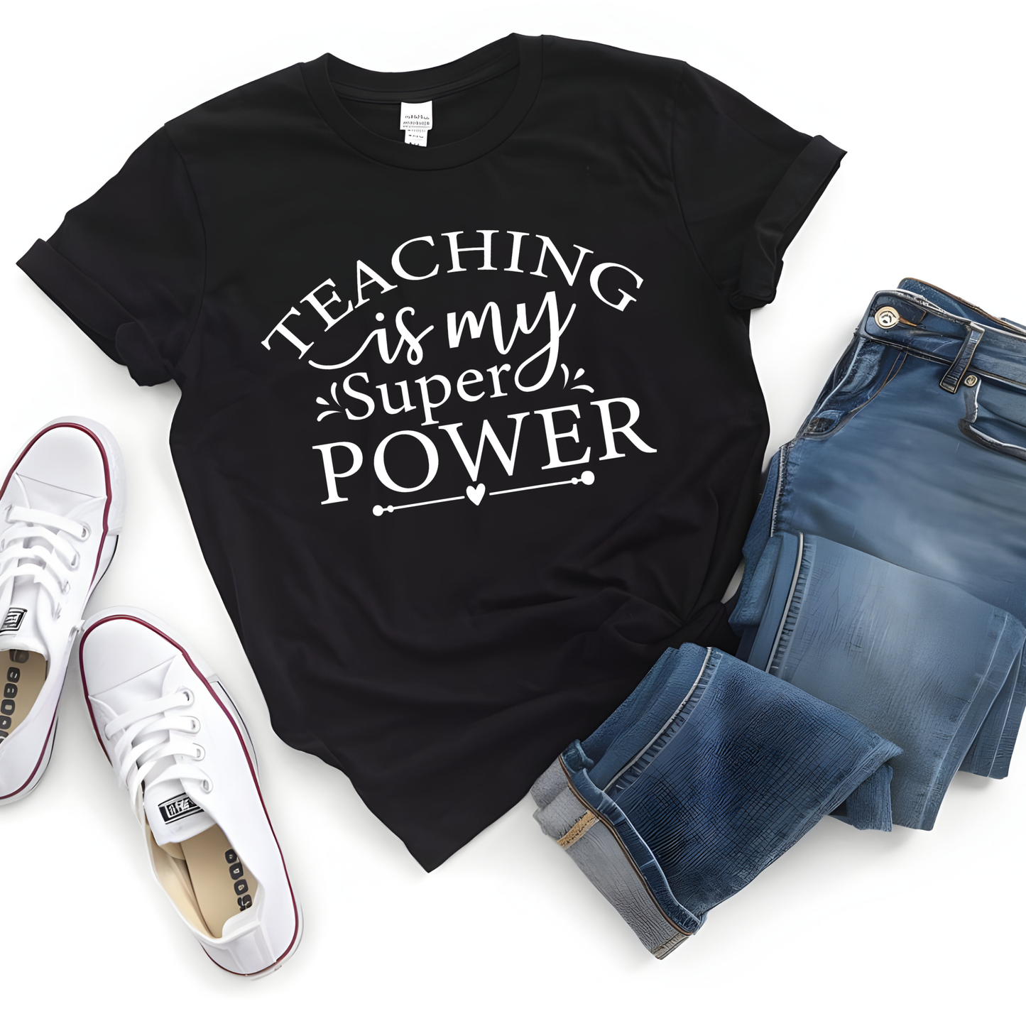 A black short-sleeve graphic T-shirt with white text reading 'TEACHING is my Super POWER' is styled in a flat lay with blue jeans and white sneakers on a white background.
