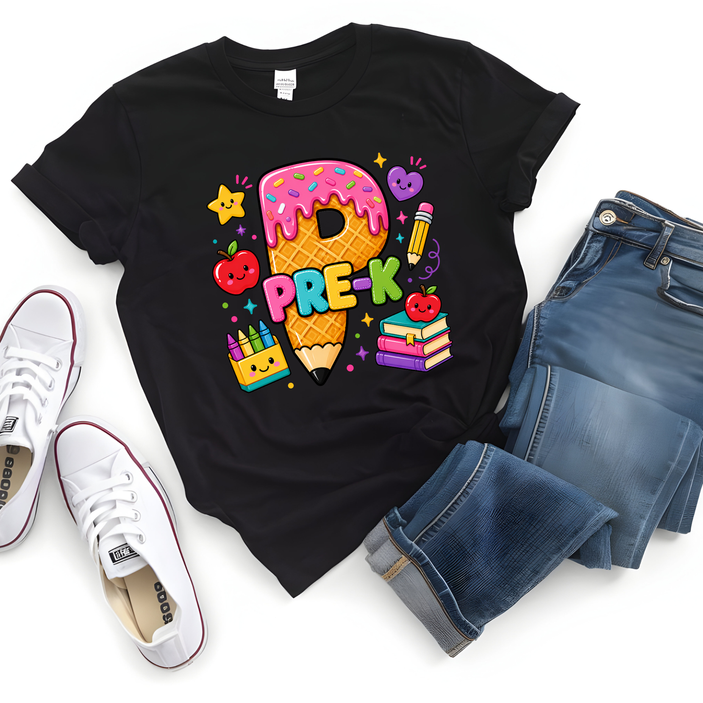 A black graphic t-shirt featuring a colorful 'PRE-K' school-themed design with a donut, pencil, stars, and books, styled in a flat lay with blue jeans and white sneakers on a white background.