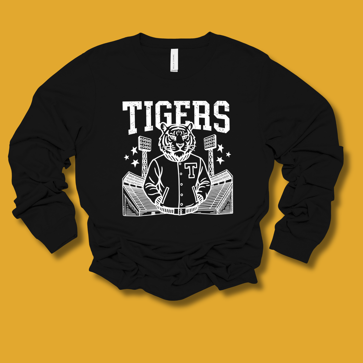 A black long-sleeve crewneck sweatshirt with a white graphic print of a tiger mascot wearing a varsity jacket, standing in front of a stadium, with the word 'TIGERS' above. The sweatshirt is laid flat on a solid mustard yellow background.