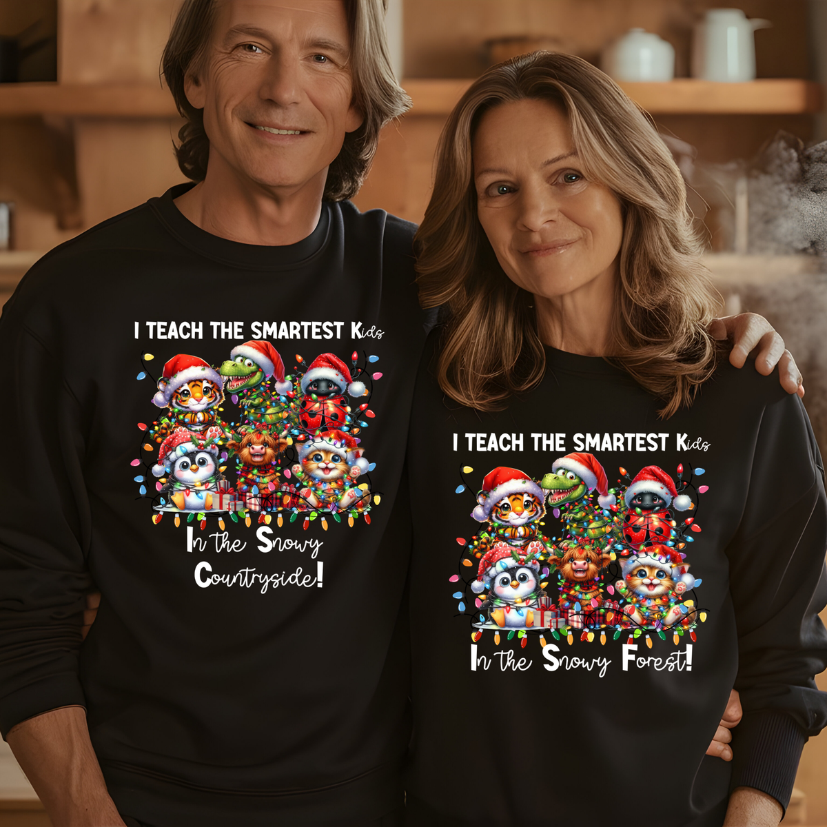 A man and a woman wearing matching black long-sleeve crewneck sweatshirts with a colorful Christmas cartoon graphic and the text 'I TEACH THE SMARTEST Kids In the Snowy Countryside! / In the Snowy Forest!'. The man wears 'Countryside' and the woman wears 'Forest' in the graphic text.