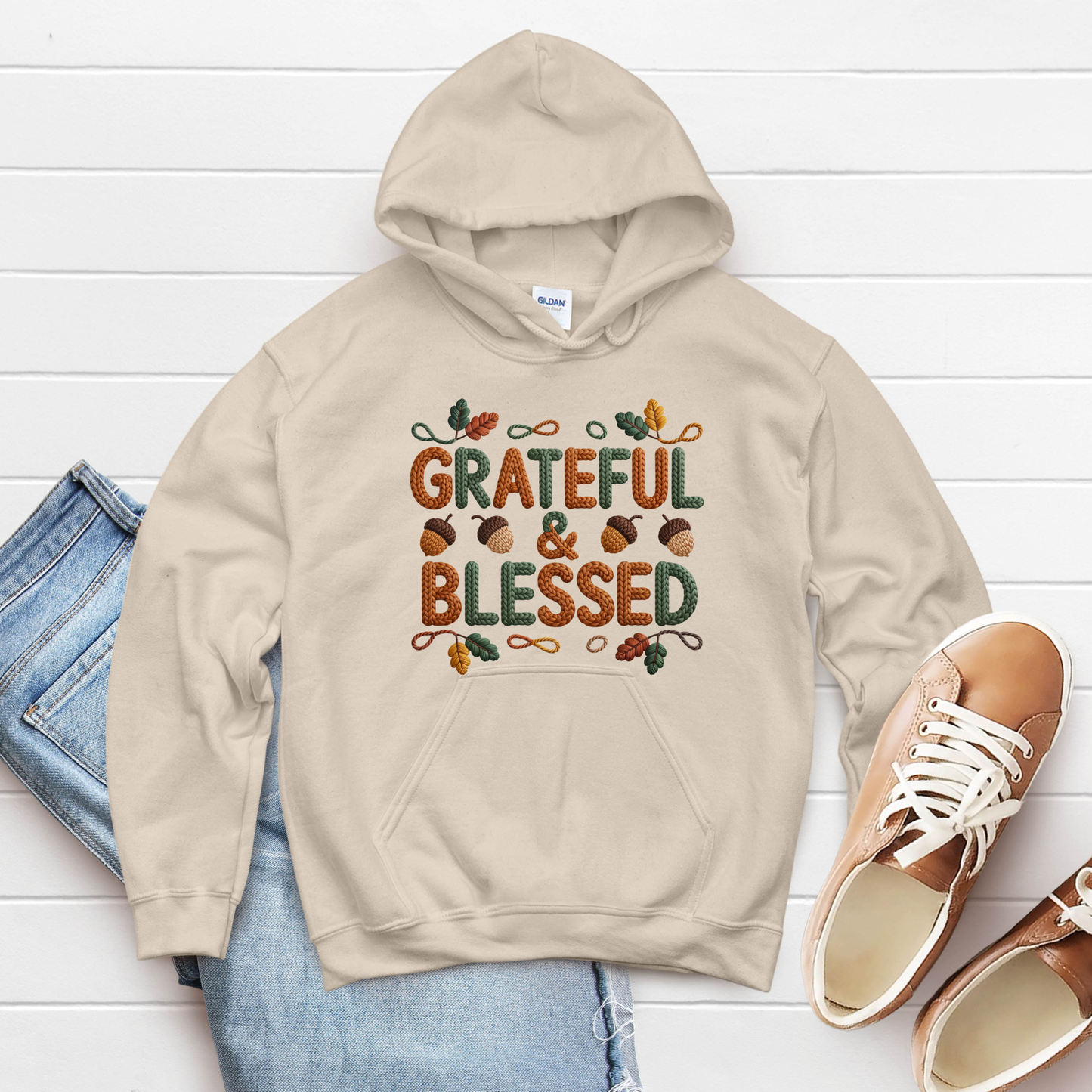 Beige hoodie with 'Grateful & Blessed' text on a white wooden surface with jeans and shoes.