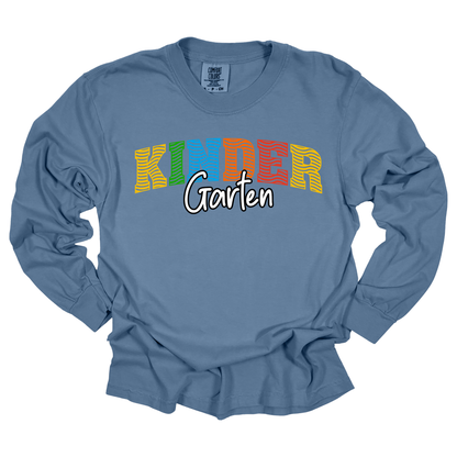 A dusty blue long-sleeve t-shirt with a graphic reading 'KINDER Garten' on the chest. The word 'KINDER' is styled in a wavy, striped multicolor font (yellow, green, orange, red), while 'Garten' is written below in a white cursive font with a black outline.