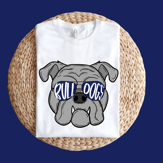 A folded white t-shirt with a cartoon bulldog graphic wearing blue sunglasses that spell 'BULL DOGS', displayed on a round woven straw mat against a dark blue background.