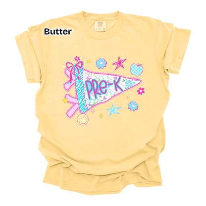A pale butter-yellow short-sleeve t-shirt with a colorful graphic of a pennant flag reading 'PRE-K' decorated with a ribbon, stars, flowers, and a donut. The word 'Butter' is printed in black text in the upper left corner.