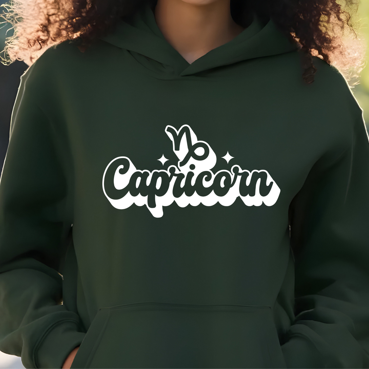 A person wearing a dark green hoodie with a white 'Capricorn' graphic and zodiac symbol printed on the chest, shown in a close-up outdoor shot.