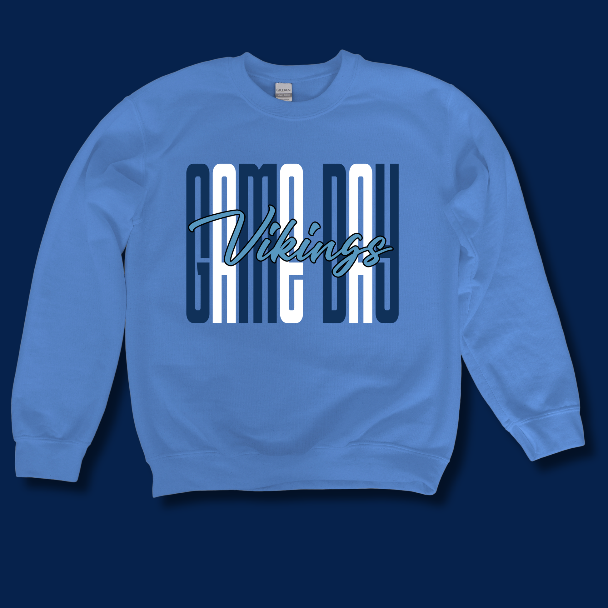 A light blue crewneck sweatshirt featuring a 'GAME DAY Vikings' graphic on the chest, displayed against a solid dark navy blue background.