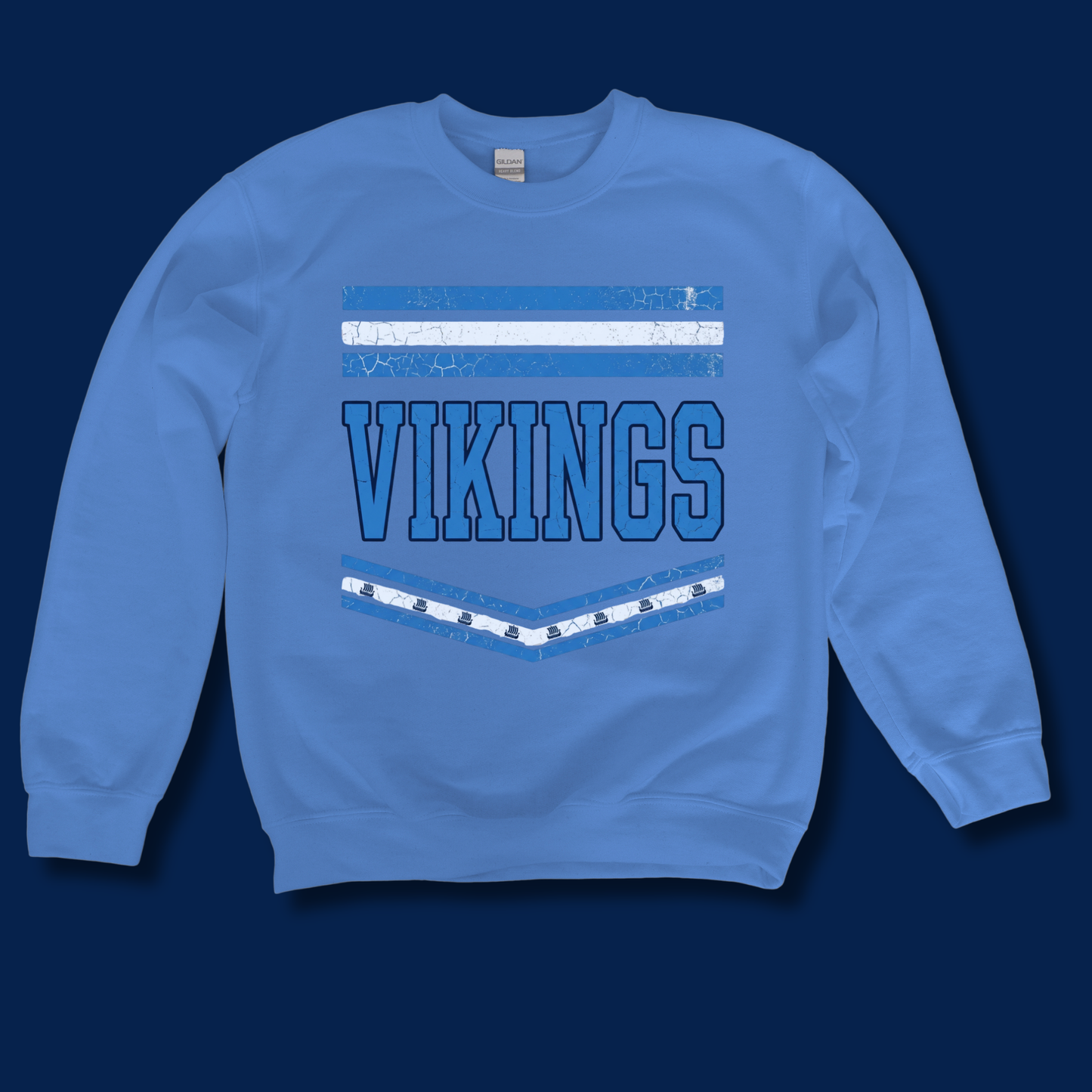A light blue crewneck sweatshirt featuring a distressed graphic on the front with the word 'VIKINGS' in blue block letters, flanked by white and blue horizontal stripes, displayed against a dark navy background.