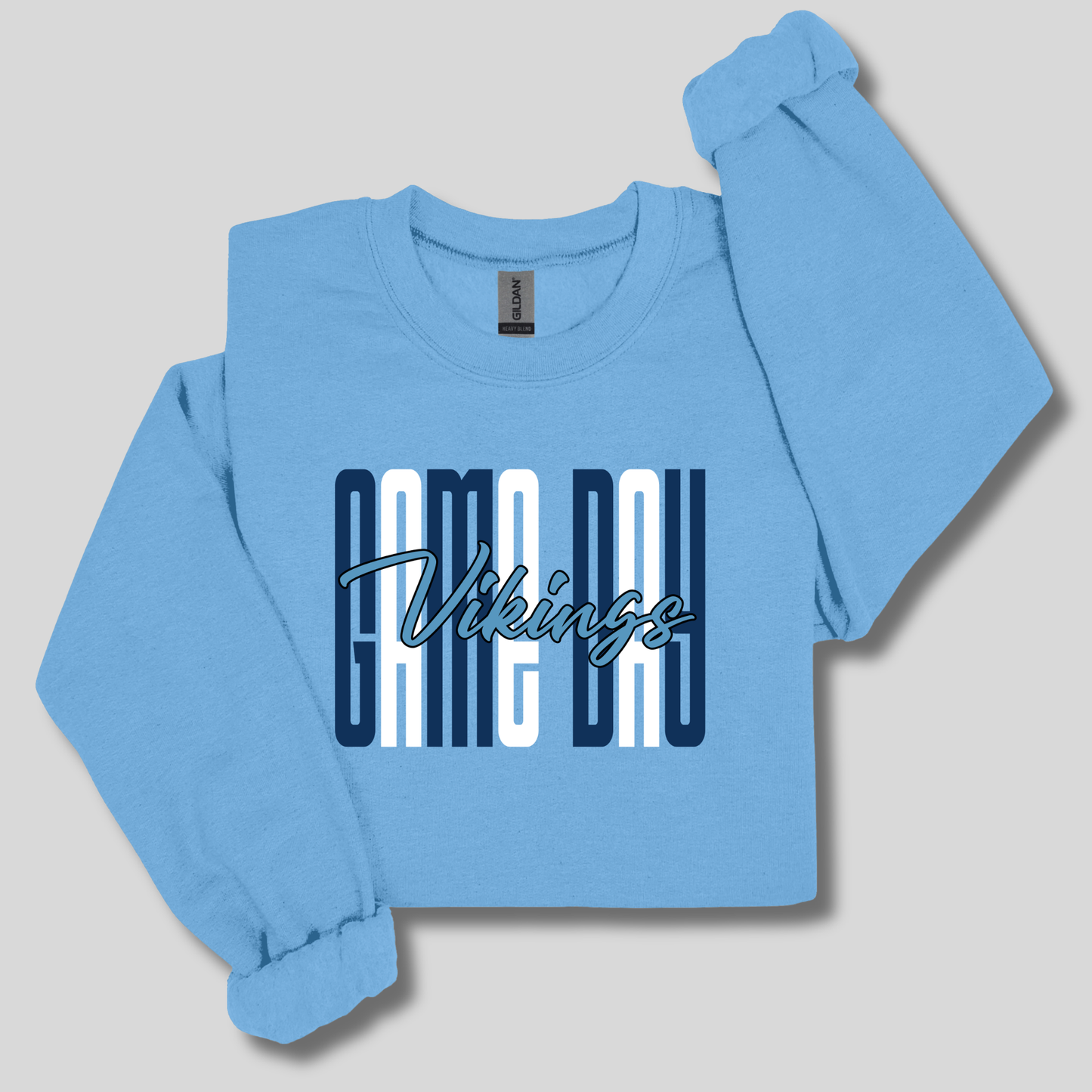 A light blue crewneck sweatshirt featuring a distressed graphic on the front with the word 'VIKINGS' in blue block letters, flanked by white and blue horizontal stripes, displayed against a dark navy background.