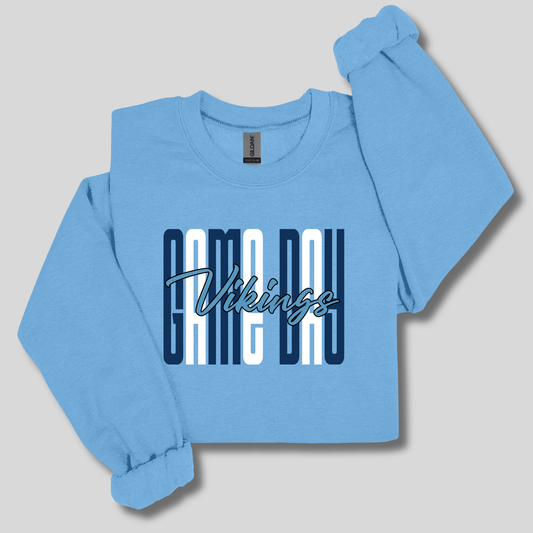A light blue crewneck sweatshirt featuring a distressed graphic on the front with the word 'VIKINGS' in blue block letters, flanked by white and blue horizontal stripes, displayed against a dark navy background.