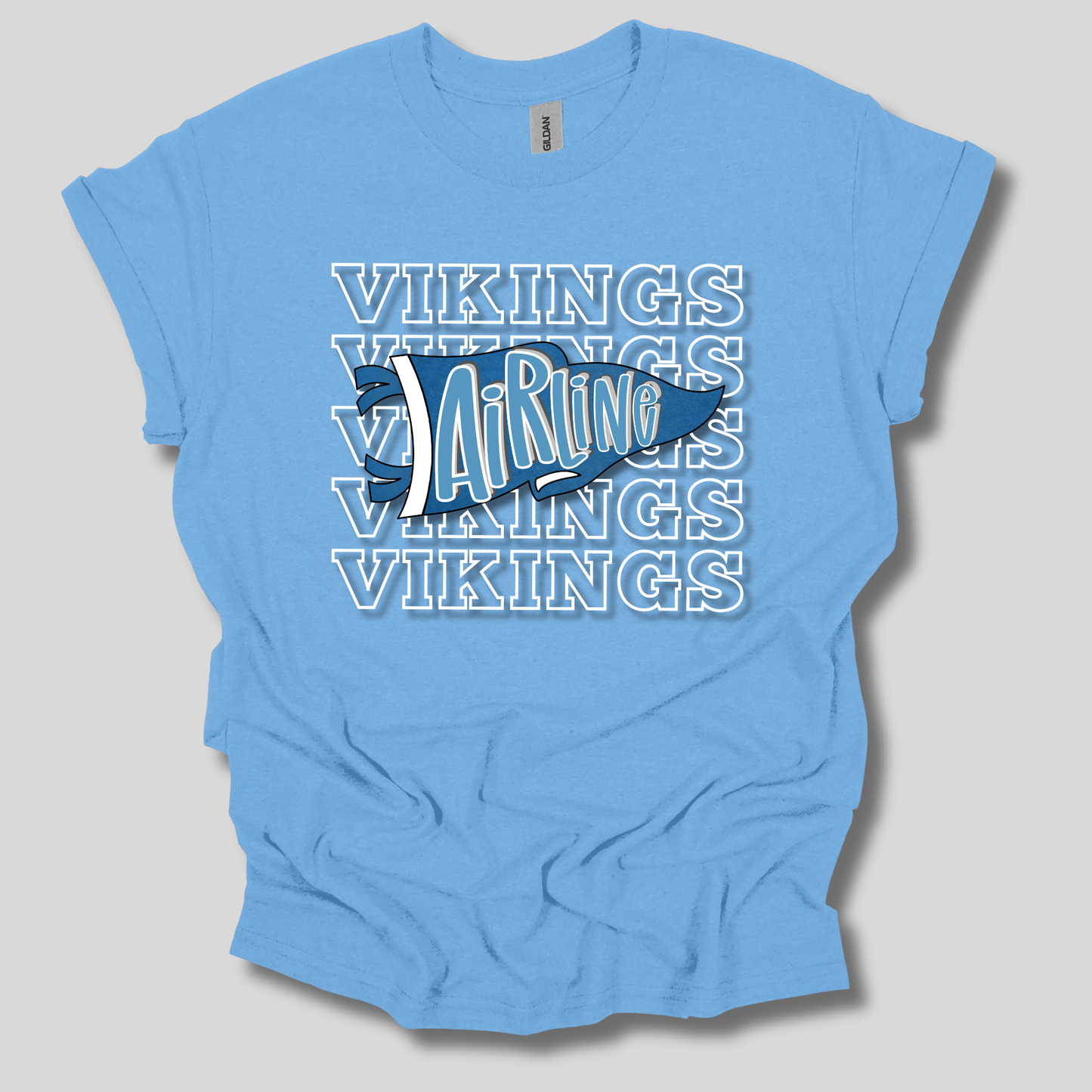 A light blue short-sleeve t-shirt with a graphic design featuring the word 'VIKINGS' repeated in white outline text, overlaid with a large blue and white Viking helmet logo in the center, displayed on a neutral gray background.