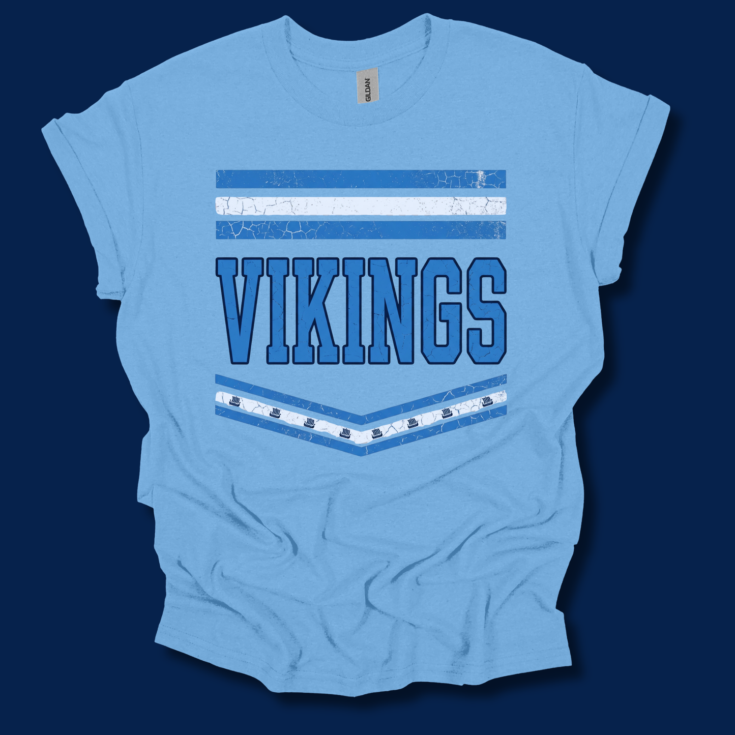 A light blue short-sleeve t-shirt with a distressed graphic featuring the word 'VIKINGS' in blue block letters, framed by blue and white horizontal stripes, laid flat against a dark navy background.