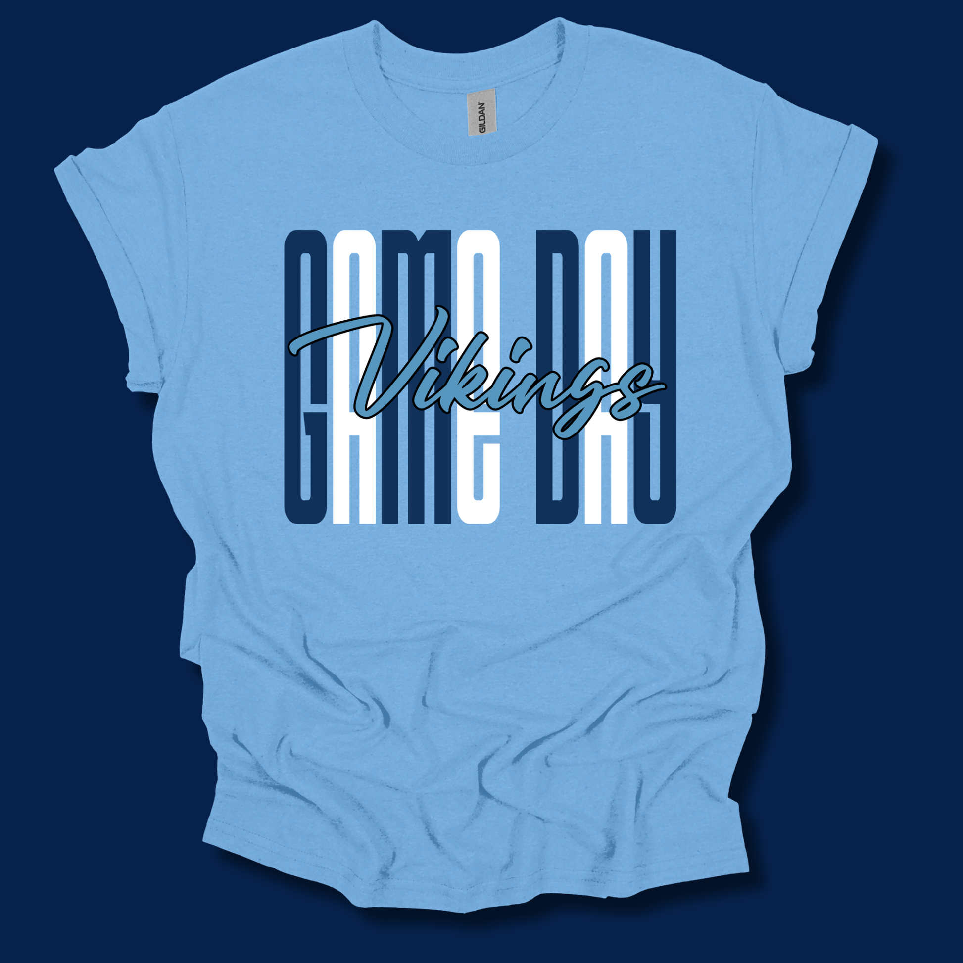 A light blue short-sleeve t-shirt with a distressed graphic featuring the word 'VIKINGS' in blue block letters, framed by blue and white horizontal stripes, laid flat against a dark navy background.