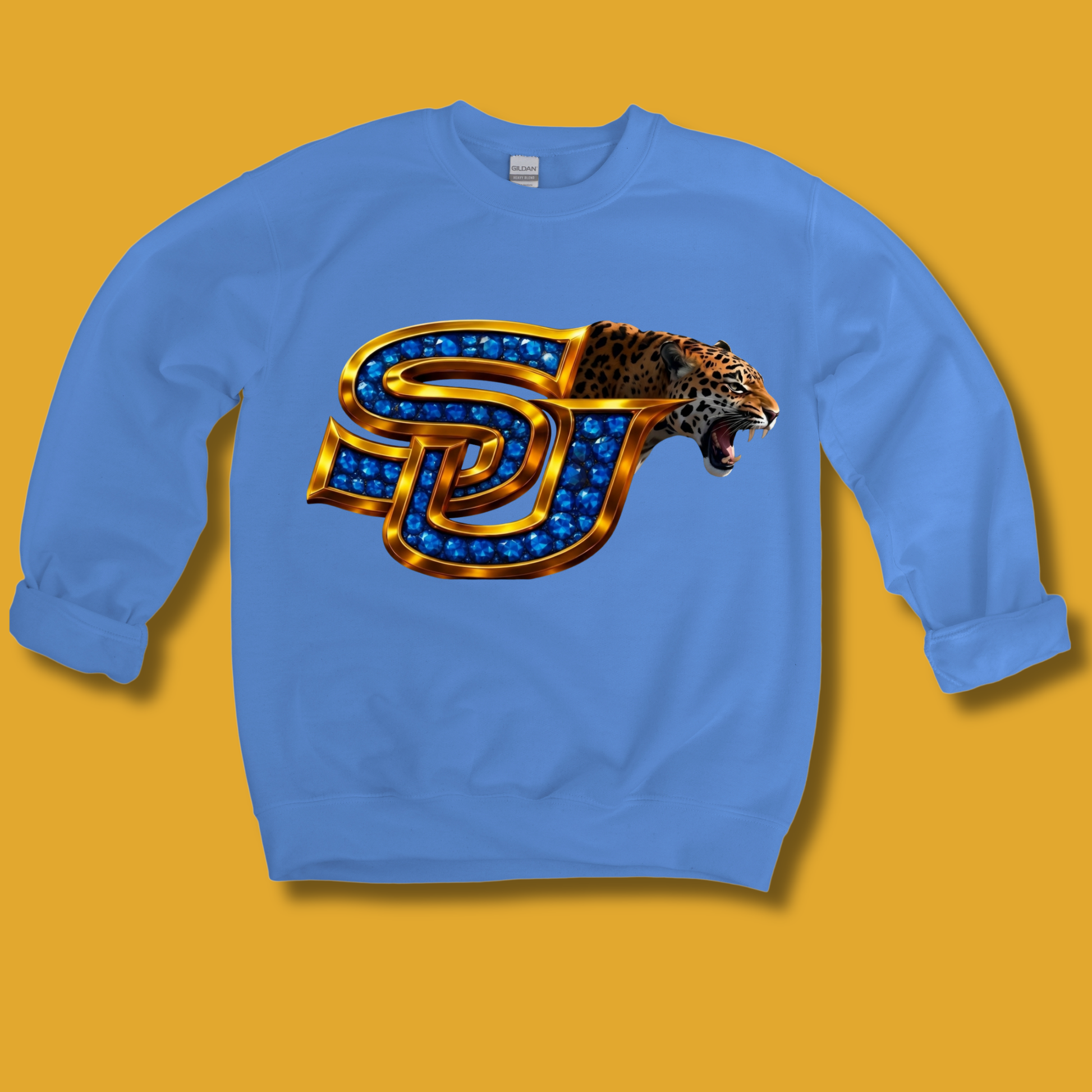 A light blue crewneck sweatshirt featuring a large graphic of the 'SJ' logo for the San Jose Sharks, styled with gold metallic lettering and blue gem-like accents, alongside a roaring leopard head. The sweatshirt is displayed flat against a solid mustard yellow background.