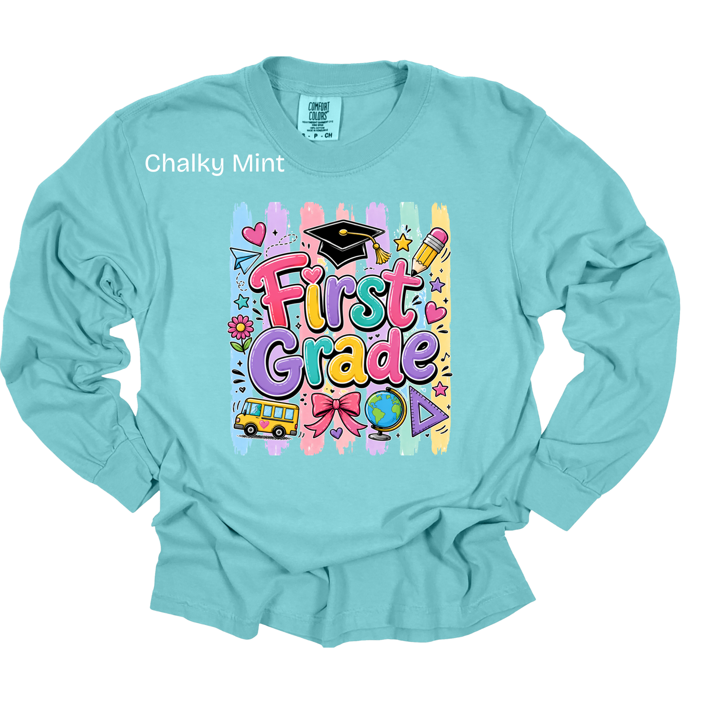 A light blue long-sleeve t-shirt with a colorful 'First Grade' graphic on the chest, featuring school-themed doodles like a graduation cap, pencil, and bus. The shirt is shown flat against a white background with a 'Chalky Mint' text overlay near the collar.