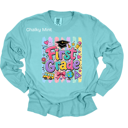 A light blue long-sleeve t-shirt with a colorful 'First Grade' graphic on the chest, featuring school-themed doodles like a graduation cap, pencil, and bus. The shirt is shown flat against a white background with a 'Chalky Mint' text overlay near the collar.