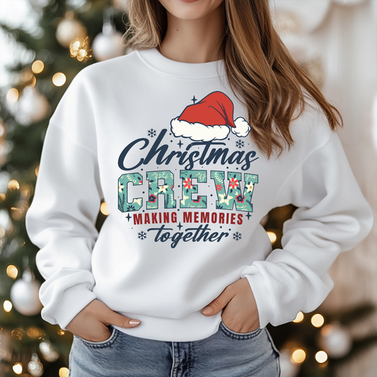 Person wearing a white sweatshirt with 'Christmas Crew Making Memories Together' text and Santa hat graphic, Christmas tree in the background.
