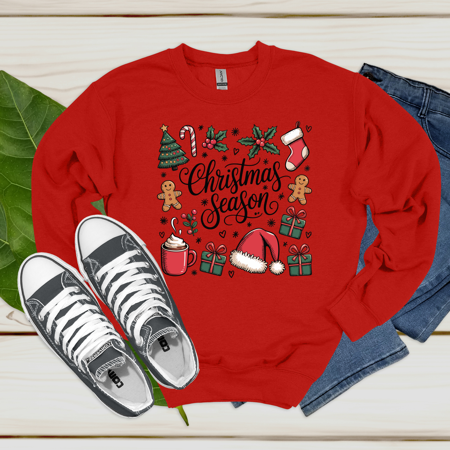 Red sweatshirt with Christmas-themed design, white sneakers, and jeans on a wooden surface.