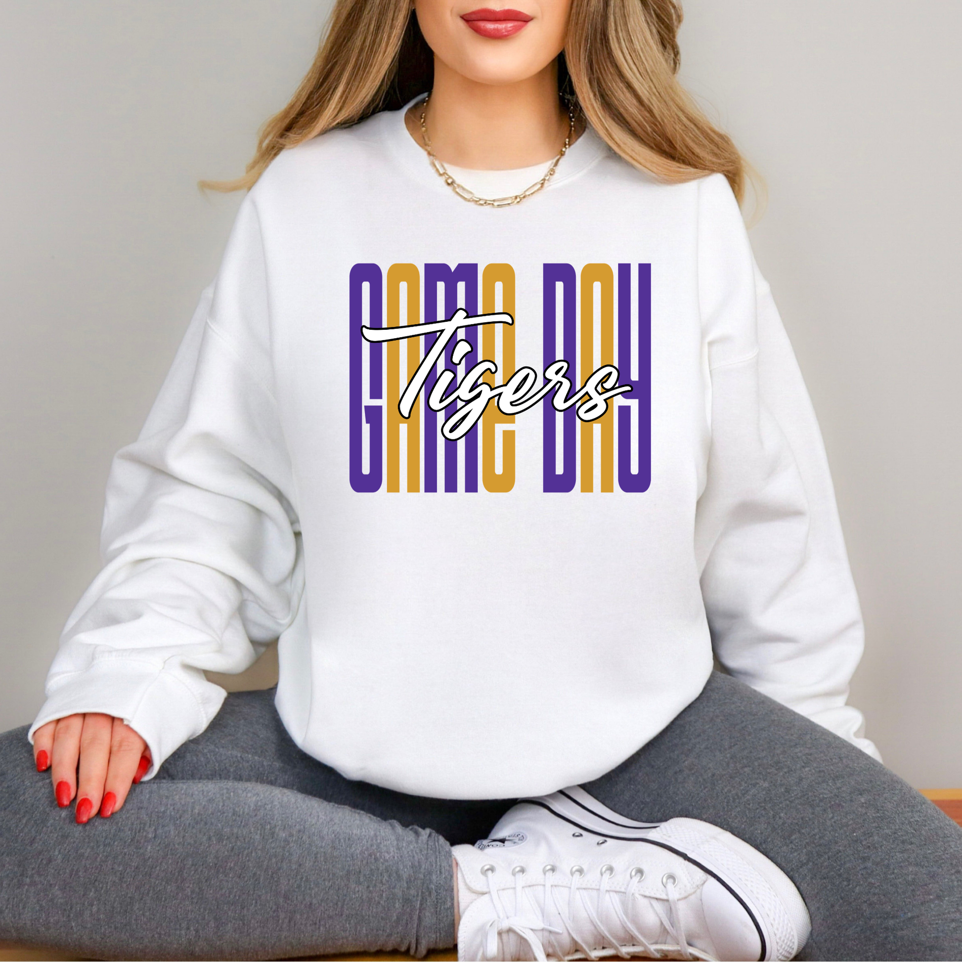 A woman wearing a white crewneck sweatshirt with a 'GAME DAY Tigers' graphic in purple and gold, paired with gray sweatpants and white sneakers.