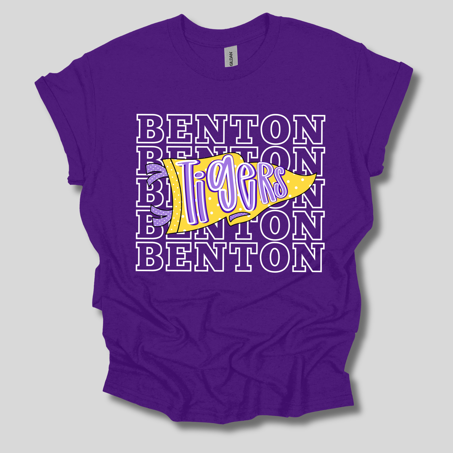 A purple short-sleeve t-shirt with a 'Benton Tigers' graphic on the front, featuring a yellow pennant with purple lettering over a background of stacked white-outlined 'BENTON' text.