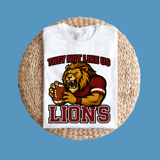 White t-shirt with lion graphic and text on a woven stool against a blue background Custom Mascot Shirt - Lions They Not Like Us Version 2 School Spirit Tee