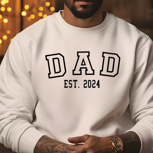 Man wearing a white sweatshirt with "DAD" in bold letters and "EST. 2024" below, featuring a heart graphic with kid names personalized on the sleeve. New Dad sweater.