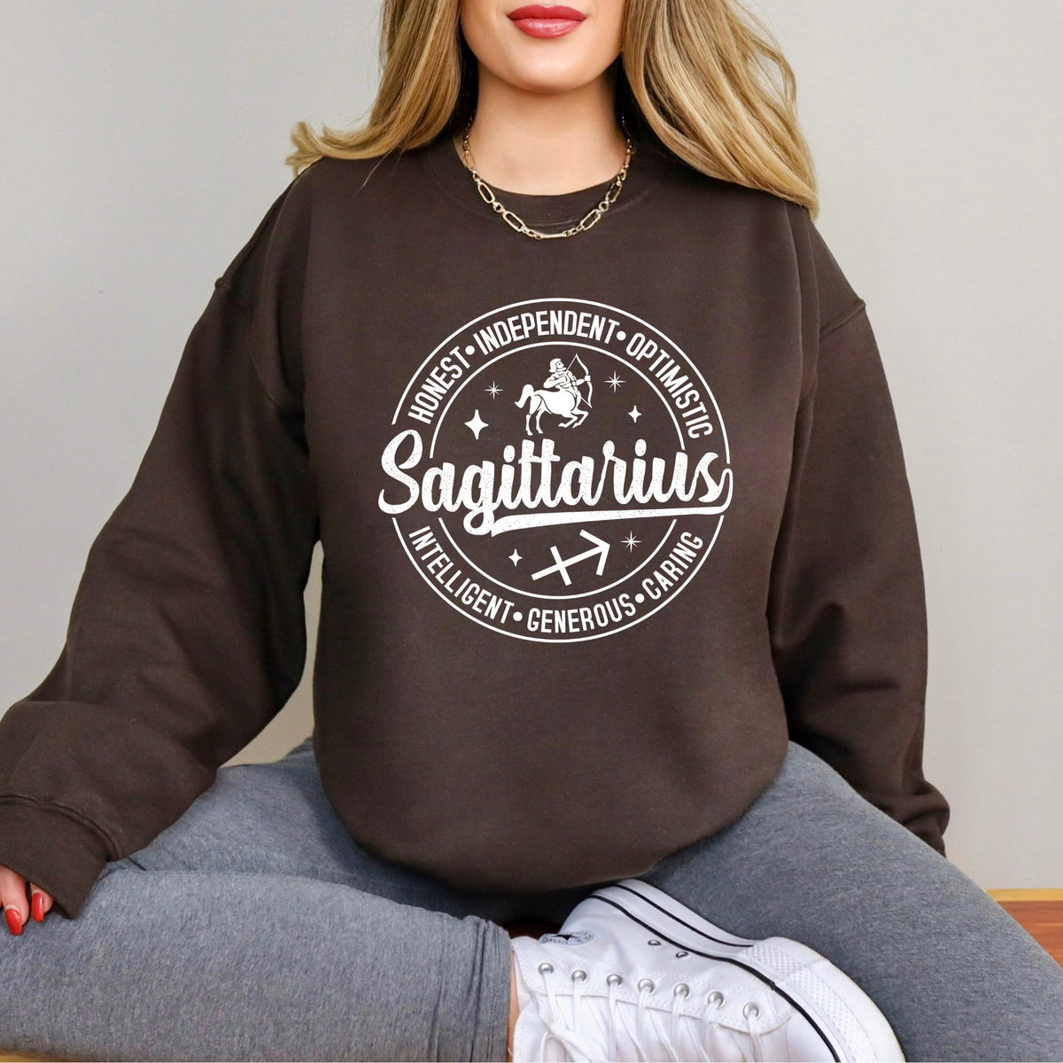 A woman wearing a dark brown crewneck sweatshirt with a white graphic print of the Scorpio zodiac sign and its qualities, paired with gray sweatpants and white sneakers.