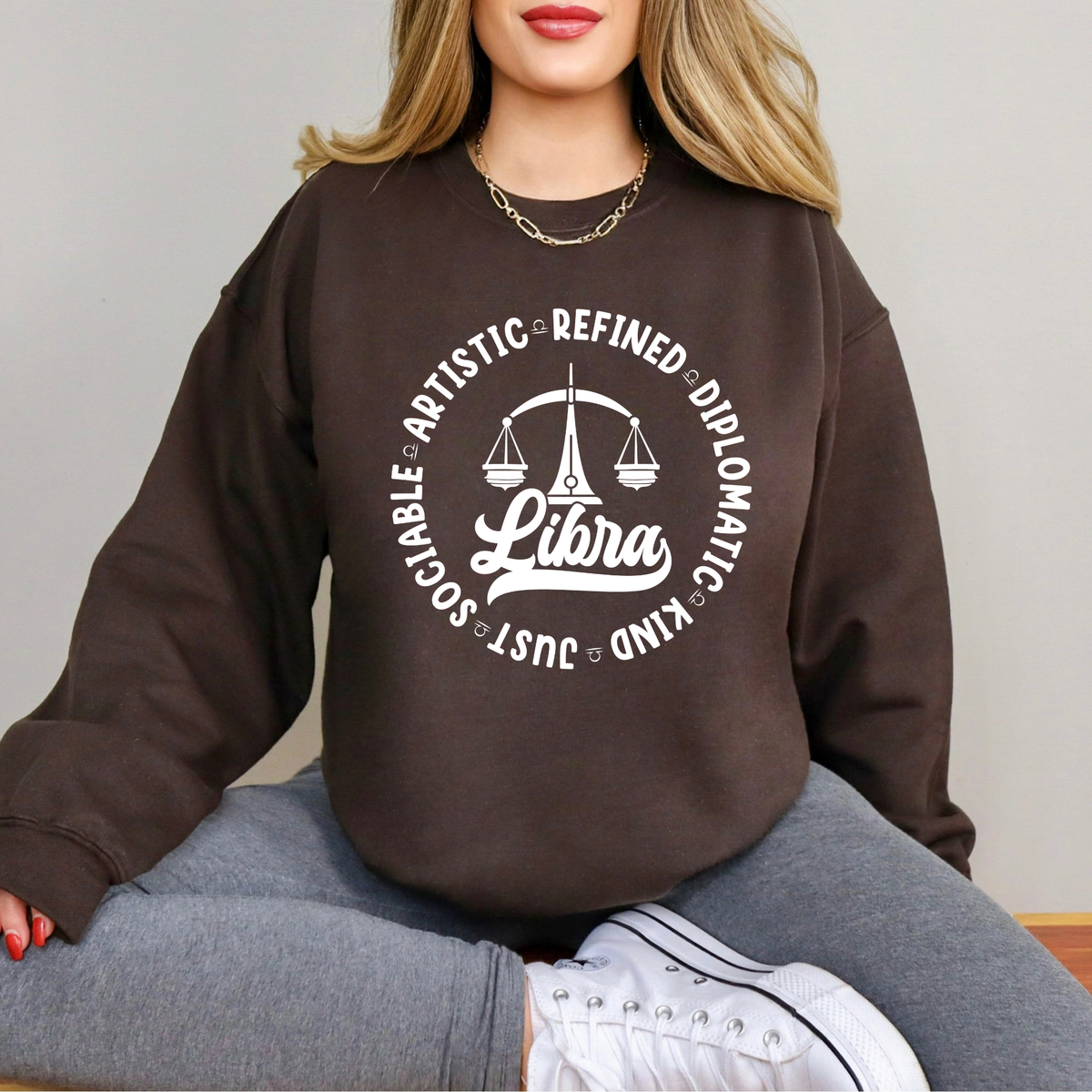 Woman wearing a white crewneck t-shirt featuring a black circular Libra zodiac graphic with a scales illustration, the word "Libra," and trait keywords like artistic, refined, diplomatic, kind, just, and sociable.