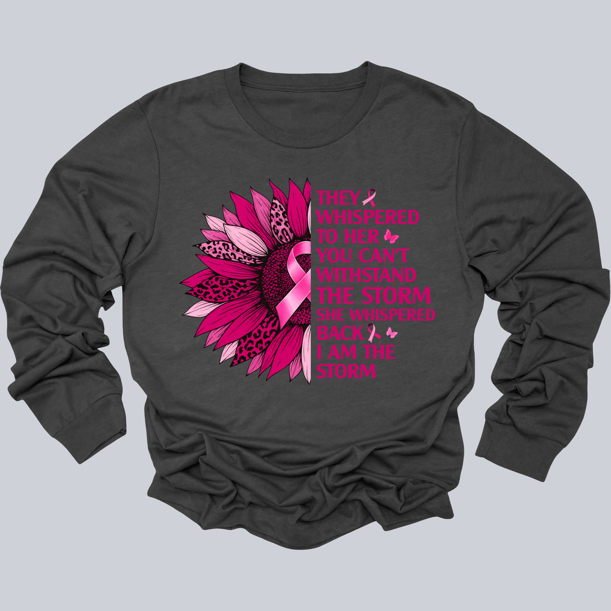 A dark charcoal gray long-sleeve t-shirt with a graphic on the front. The graphic features a stylized pink and leopard-print flower alongside pink text that reads, 'THEY WHISPERED TO HER YOU CAN'T WITHSTAND THE STORM SHE WHISPERED BACK I AM THE STORM' decorated with small pink butterflies.