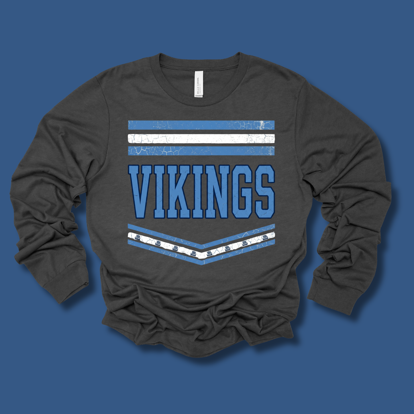A dark charcoal gray long-sleeve t-shirt with a distressed blue and white graphic on the front reading 'VIKINGS' with horizontal stripes, laid flat on a solid blue background.