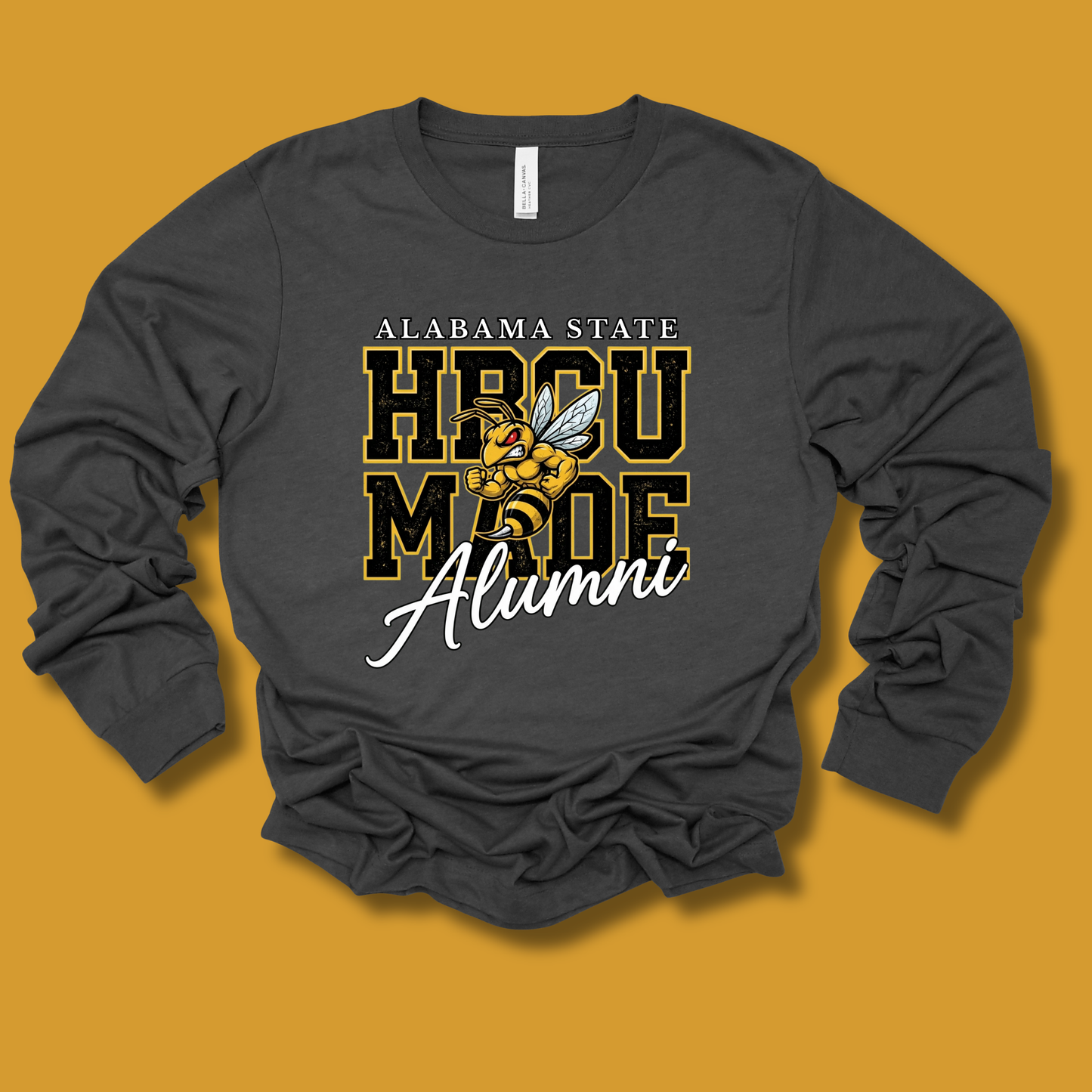 A dark gray long-sleeve t-shirt with a graphic print on the front, laid flat against a solid mustard yellow background. The graphic features the text 'ALABAMA STATE', 'HPOU MADE', and 'Alumni' in white and gold, along with the Alabama State Hornets mascot.