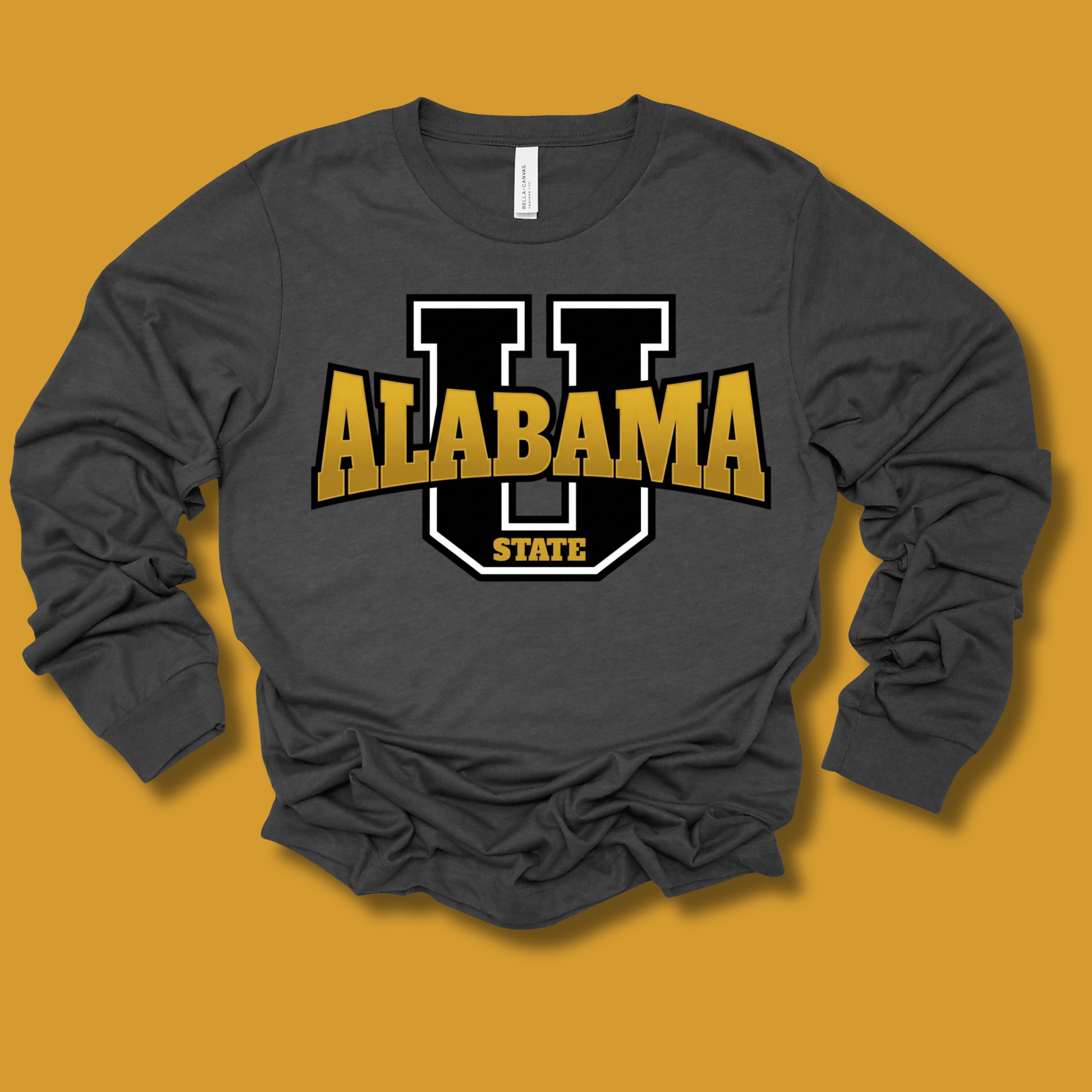 A dark gray long-sleeve t-shirt with a graphic of a large black and white letter 'U' outlined in gold, with 'ALABAMA STATE' printed in gold and white. The shirt is laid flat on a solid mustard yellow background.