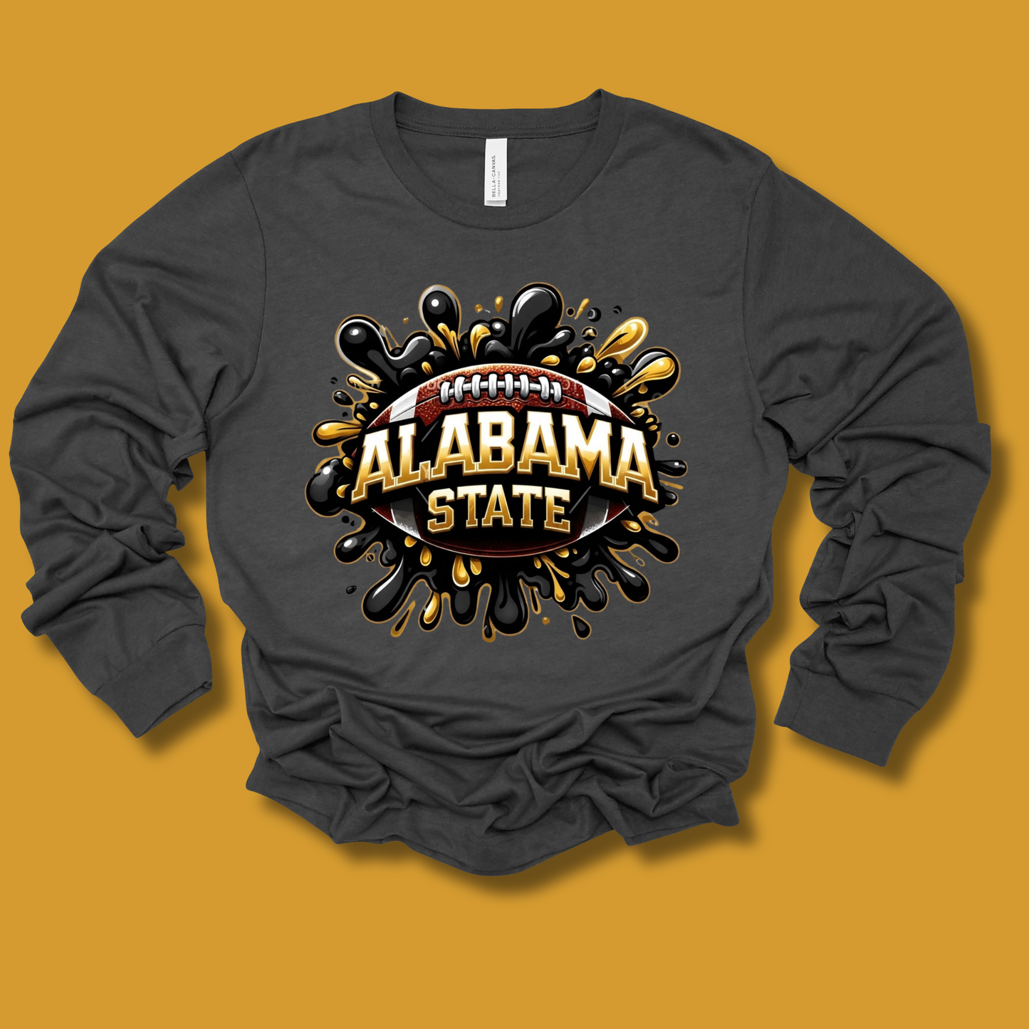 A dark gray long-sleeve t-shirt with a graphic design on the front featuring a football and the words 'ALABAMA STATE' in gold and white, surrounded by black and gold paint splatters, displayed on a solid mustard yellow background.