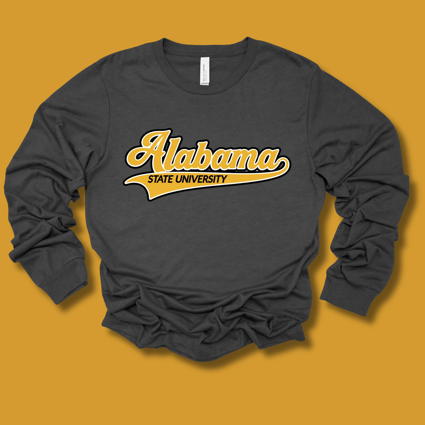 A dark charcoal gray long-sleeve t-shirt with 'Alabama State University' printed in yellow and white across the chest, laid flat on a solid mustard yellow background.