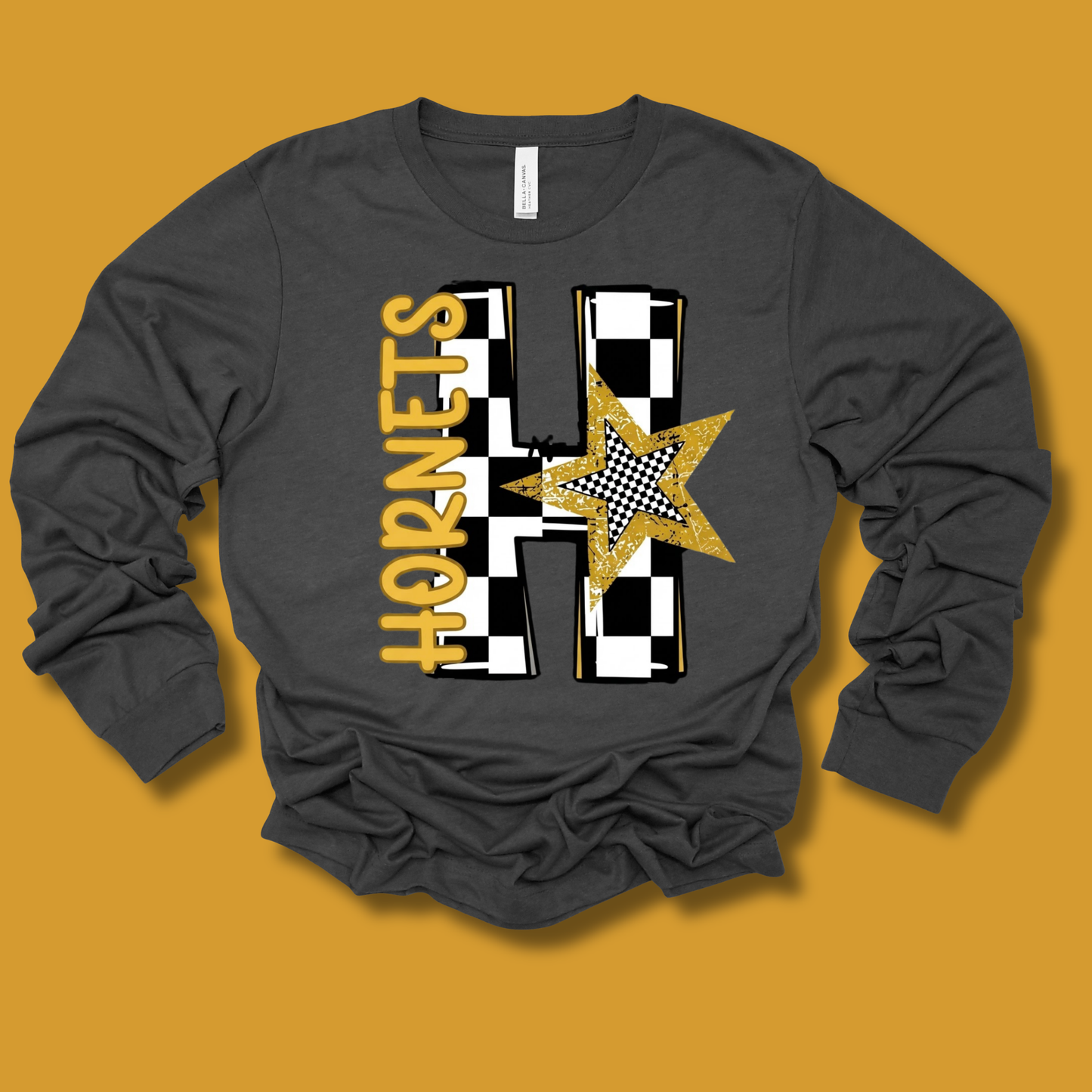 A dark gray long-sleeve t-shirt with a custom graphic design featuring the word 'HORNETS' in gold, a large black-and-white checkered 'H', and a gold glitter star, laid flat on a mustard yellow background.