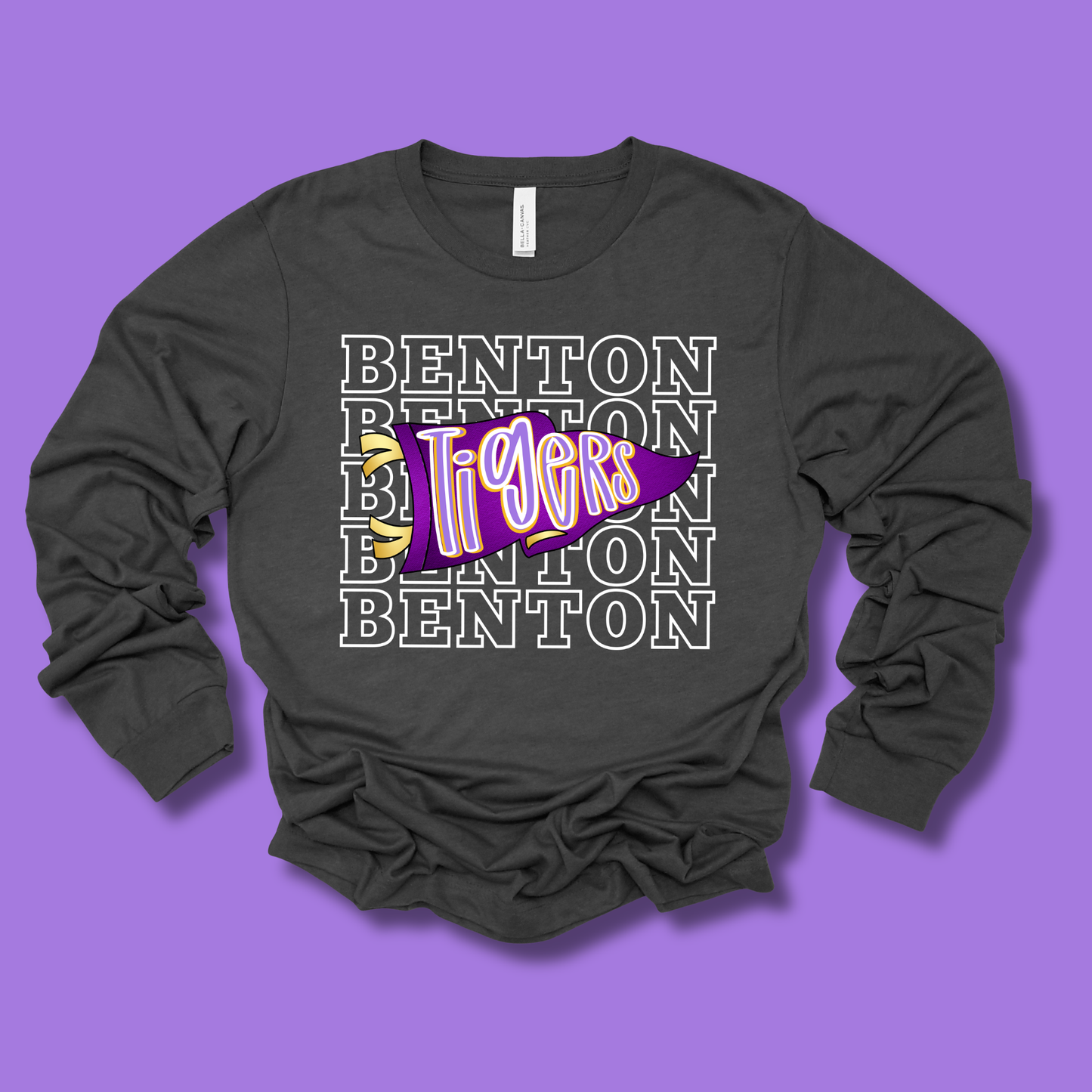 A dark charcoal gray long-sleeve t-shirt with a graphic design on the front, laid flat against a solid purple background. The design features the word 'BENTON' repeated in white outline text, overlaid with a purple and pink stylized 'Tigers' graphic.