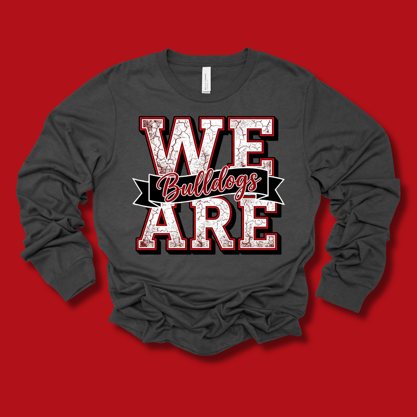A dark gray long-sleeve t-shirt with a distressed graphic reading 'WE ARE Bulldogs' in red, white, and black, displayed on a solid red background.
